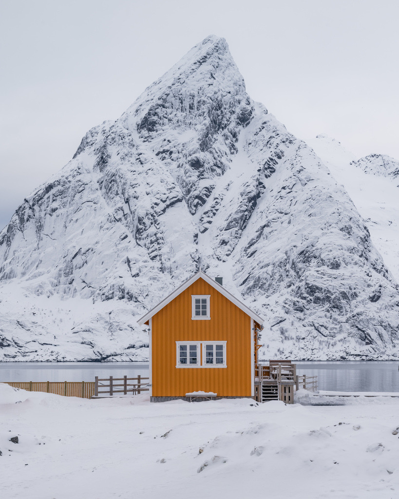 Gelbes Haus vor norwegischer Bergkulisse | Ein einzelnes gelbes Haus steht vor einem mächtigen, verschneiten Berg. Der starke Farbkontrast und die reduzierte Winterlandschaft machen das Motiv zu einer klaren, grafischen Komposition. - Realisiert mit Pictrs.com