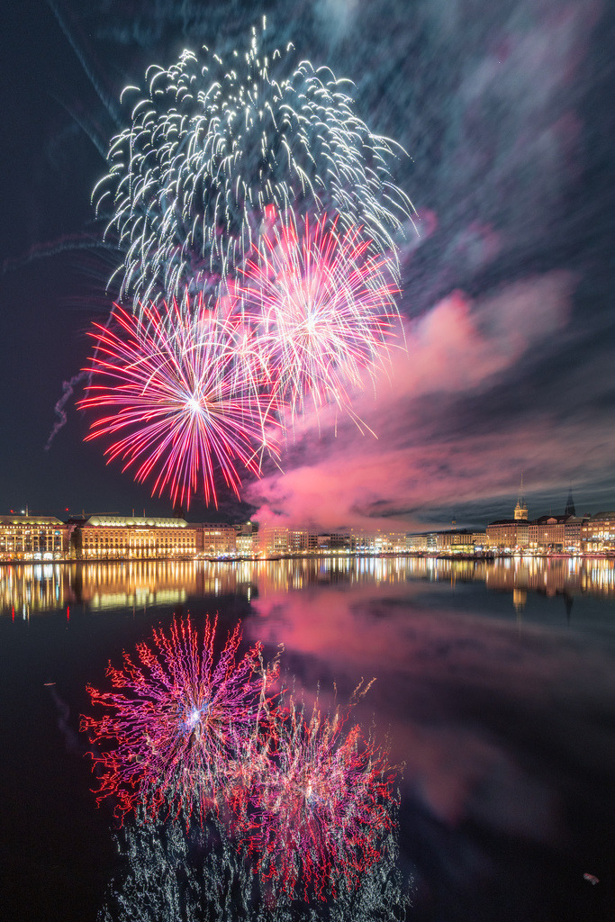 Feuerwerk in Rot und Weiß über Hamburg | Rote und weiße Feuerwerkskörper leuchten über der Hamburger Alster und spiegeln sich klar im Wasser. Rauch und Wolken verleihen der Aufnahme eine besondere Atmosphäre. - Realisiert mit Pictrs.com