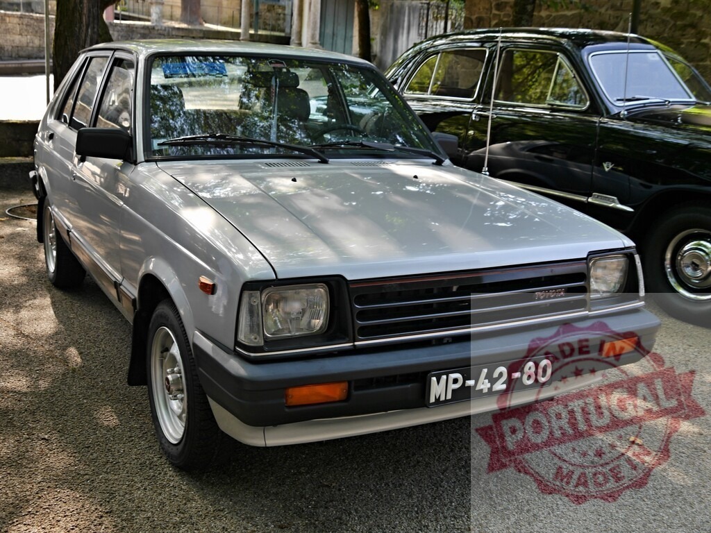 Toyota Starlet hatchback vintage car parked outdoors | Toyota Starlet, a silver vintage hatchback, parked on gravel next to an older dark car - Realized with Pictrs.com