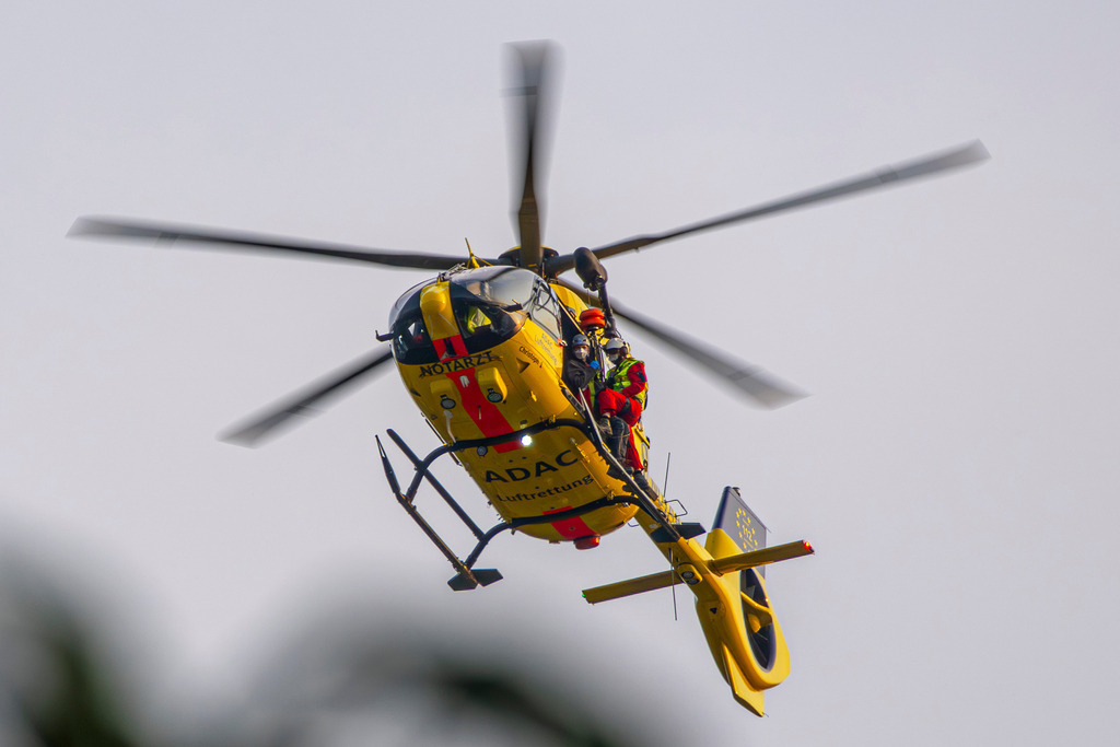 Bergretter | Ein markanter gelber ADAC-Rettungshubschrauber (Christoph 1) befindet sich im Schwebeflug vor einem klaren, hellen Himmel. An der Seite ist die Rettungswinde im Einsatz: Ein Luftretter sichert eine Person, während sie gemeinsam am Seil unterhalb der offenen Kabinentür hängen – ein intensiver Einblick in die lebensrettende Arbeit der Bergwacht oder Luftrettung. - Realisiert mit Pictrs.com