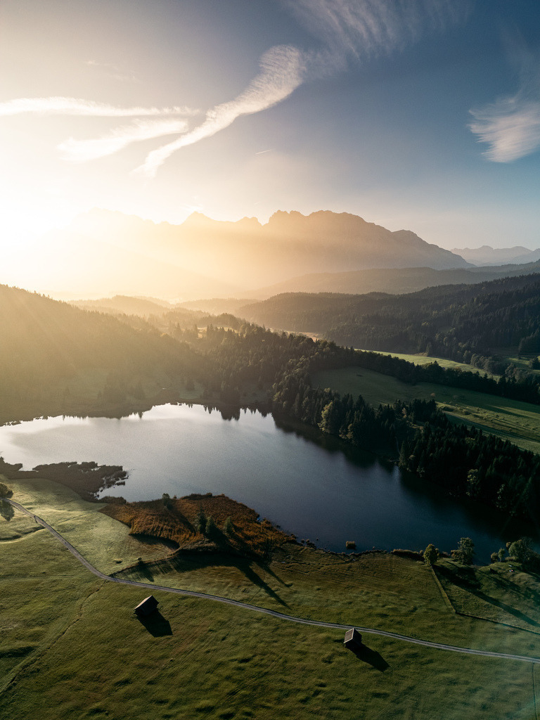 Bergsee im goldenen Morgenlicht | Ein stiller Bergsee liegt unterhalb der Alpen und wird vom warmen Licht des frühen Morgens umgeben. Die erhöht aufgenommene Perspektive verbindet See, Wiesen und Bergsilhouette zu einer weiten Landschaftskomposition. - Realisiert mit Pictrs.com