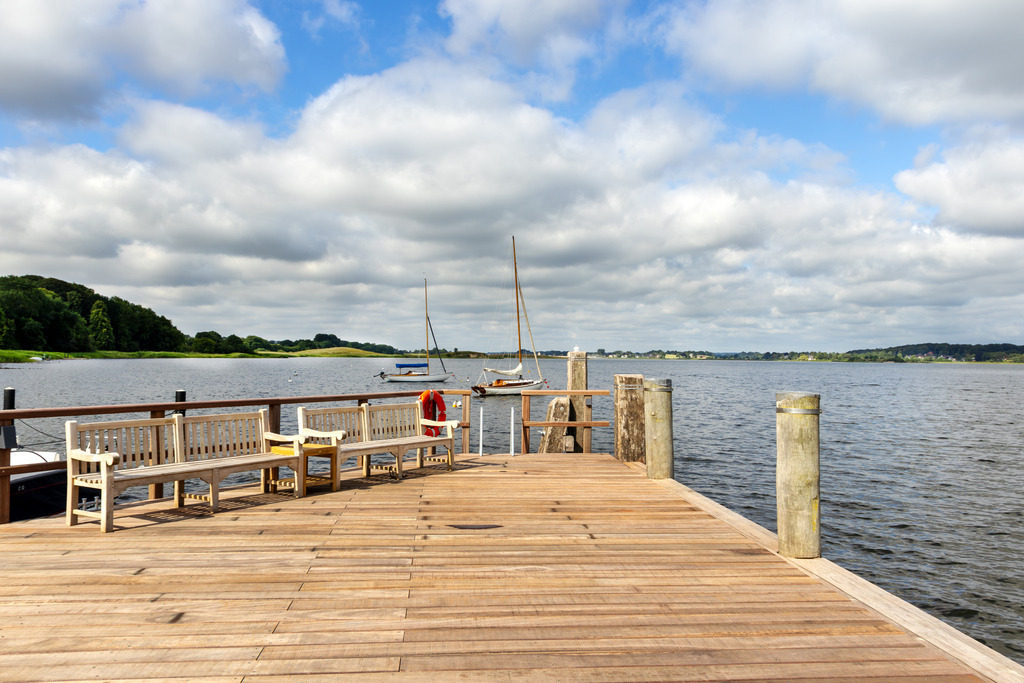 Wandbild: Steg in Sieseby an der Schlei | Dieses Wandbild im Querformat zeigt den neuen Steg in Sieseby an der Schlei. Auf dem Steg befinden sich zwei Bänke. Auf der Schlei kann man zwei Segelboote sehen. Am blauen Himmel befinden sich zahlreiche sommerliche Wolken - Realisiert mit Pictrs.com