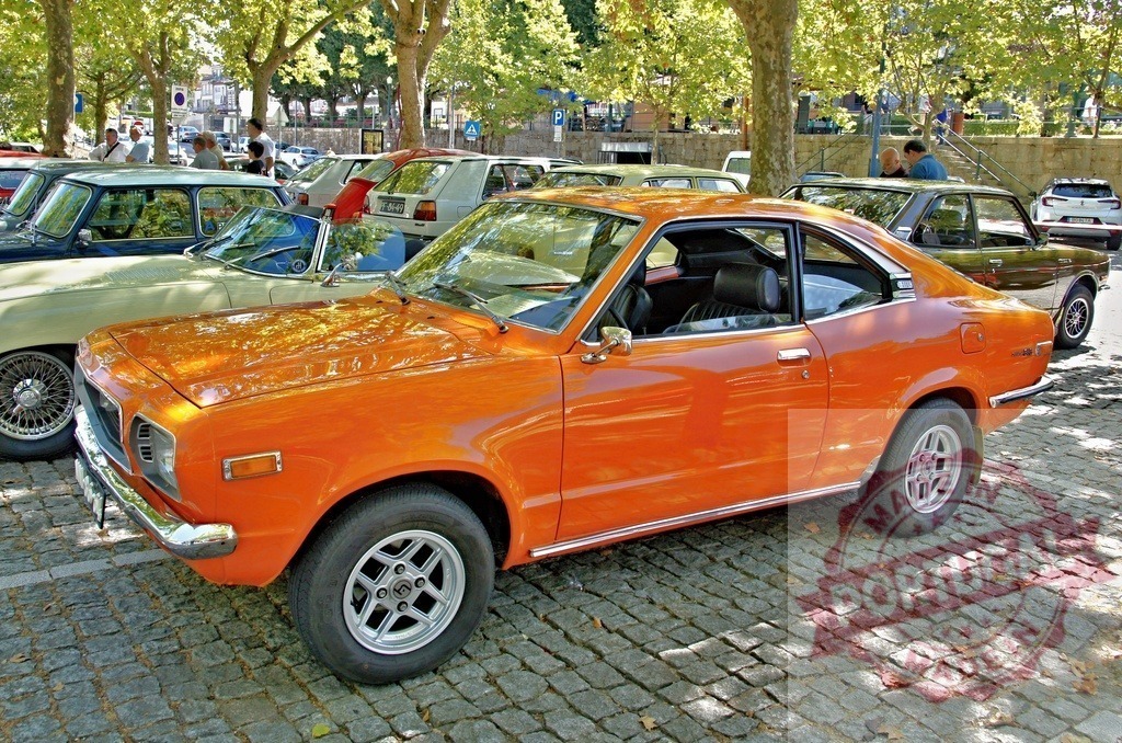 Shiny orange Mazda RX-3 coupe parked on cobblestone street | Classic orange Mazda RX-3 parked among other vintage cars on a cobblestone street under the shade of trees - Realized with Pictrs.com