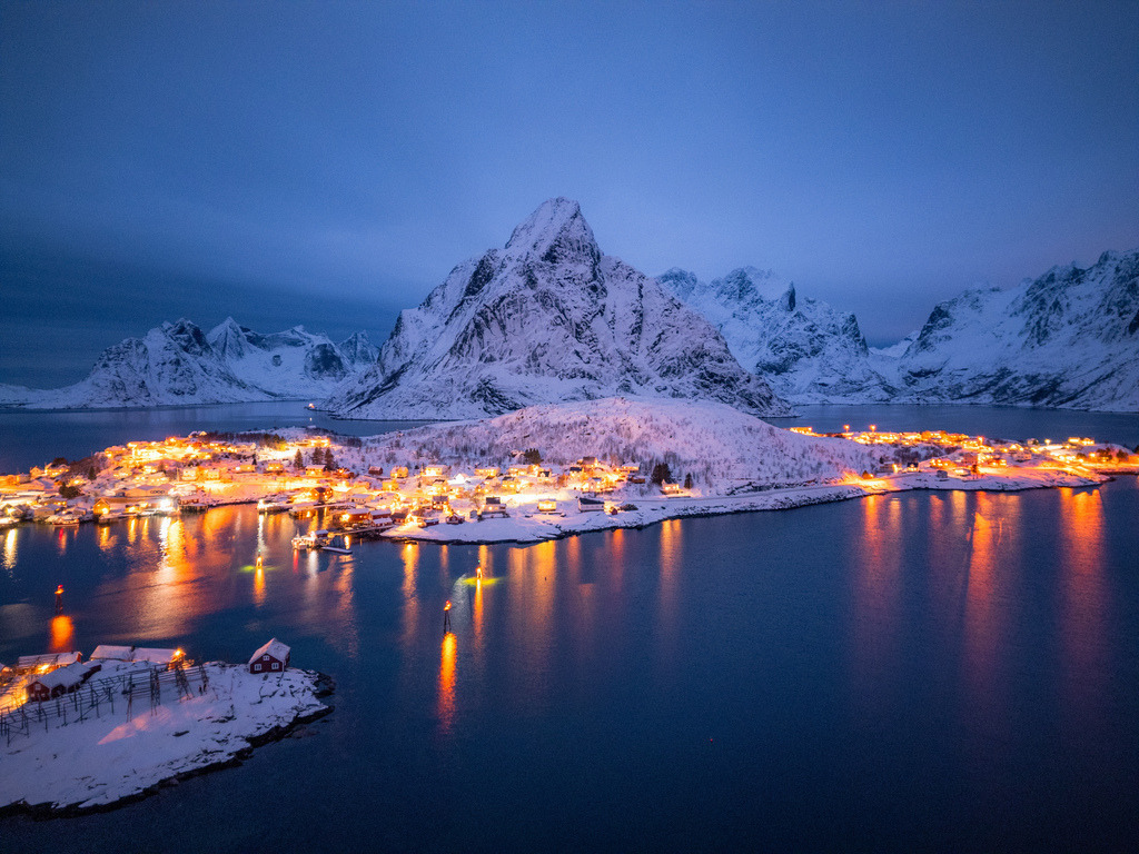 Norwegischer Küstenort zur blauen Stunde | Die warmen Lichter eines norwegischen Küstenortes spiegeln sich zur blauen Stunde im ruhigen Wasser. Im Hintergrund erhebt sich eine eindrucksvolle verschneite Berglandschaft. - Realisiert mit Pictrs.com