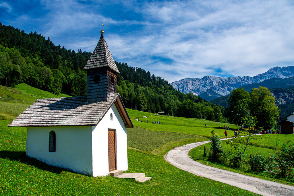 Alpine Ruhe an der Kapelle in Graseck | Eine kleine, weiß getünchte Kapelle mit traditionellem Holzschindeldach liegt friedlich eingebettet in saftig grüne Almwiesen auf dem Hochplateau Graseck. Ein schmaler Weg schlängelt sich durch die bayerische Kulturlandschaft nahe Garmisch-Partenkirchen, während im Hintergrund die imposante Kulisse des Wettersteingebirges emporragt. Unter dem leicht bewölkten Sommerhimmel vermittelt dieses Motiv ein Gefühl von zeitloser Ruhe, Besonnenheit und der idyllischen Harmonie zwischen Mensch und Natur in den Alpen. - Realisiert mit Pictrs.com