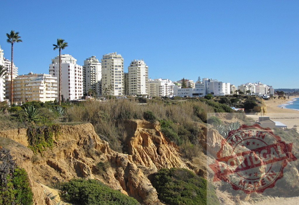 Praia da Rocha coastline with cliffs and city buildings. Algarve - Silves | Algarve coastline view showing sandy beach, ocean, and cityscape under a clear blue sky - Realized with Pictrs.com