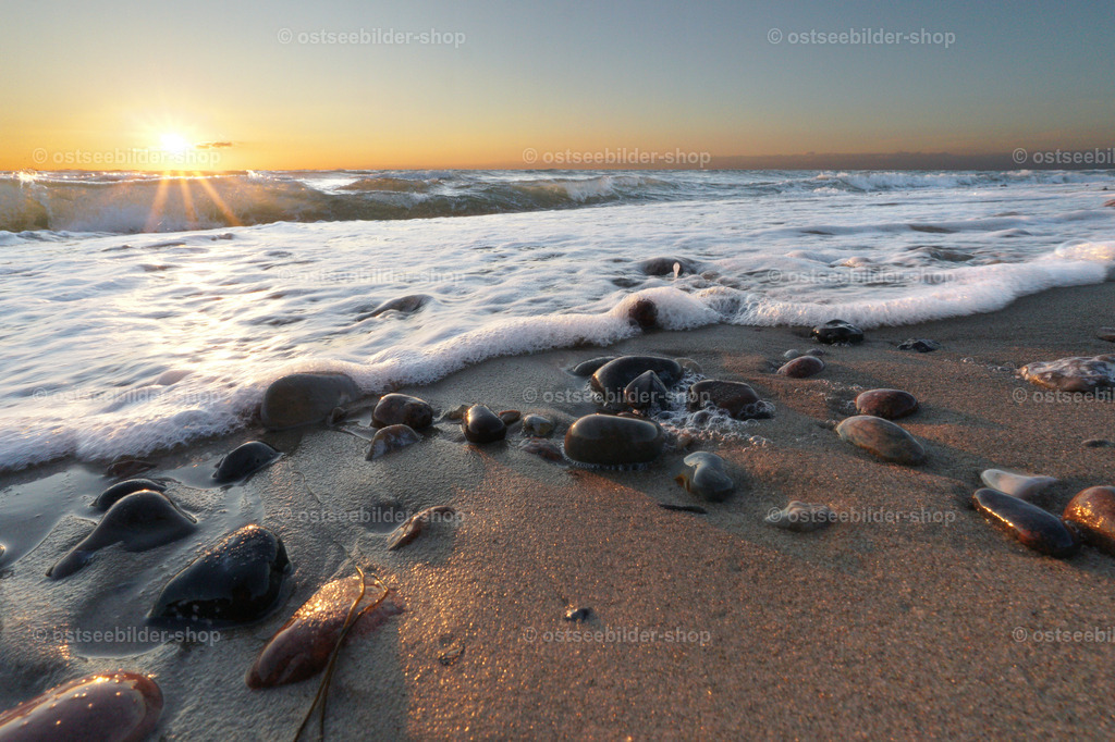 Auslaufende Brandung am Ostseestrand | Eine auslaufende Welle trifft auf Steine am Ostseestrand. Über den Wellenkämmen geht die Sonne unter.