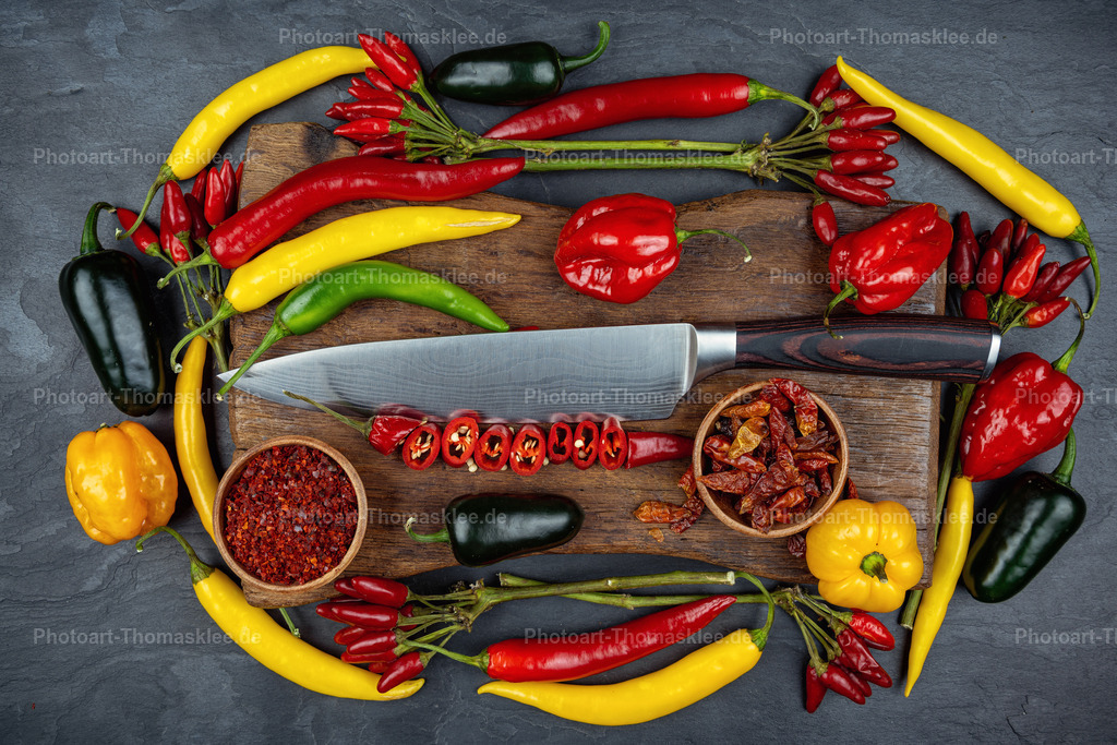 Bunte Chilis mit Messer auf Holzschneidebrett | Eine kreative Anordnung verschiedener Chilisorten bildet einen farbenfrohen Kreis um ein dunkles Holzschneidebrett. Gelbe Bananenpaprika, rote Habaneros und kleine grüne Chilis schaffen einen lebendigen Kontrast zum grauen Schieferhintergrund. Das Küchenmesser und kleine Holzschalen mit Chilipulver ergänzen die Szene perfekt. Diese durchdachte Komposition vereint frische und getrocknete Gewürze zu einem stimmungsvollen Stillleben. Die intensive Farbpalette macht Lust auf scharfe Küchenexperimente und würzige Gerichte. - Realisiert mit Pictrs.com