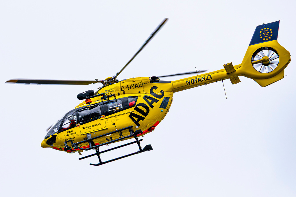 Lebensretter in Schräglage: Dynamik der ADAC-Luftrettung | Mit beeindruckender Dynamik neigt sich der ADAC-Rettungshubschrauber „Christoph Murnau“ (H145 / EC145 T2, Kennung D-HYAE) mitten im Flug vor einem strahlend hellen Hintergrund. Die Freistellung im Flug betont jedes Detail der modernen Technik – vom Fenestron-Heckrotor über die Rettungswinde bis hin zur voll besetzten Pilotenkabine. Das Bild fängt die pure Energie, Präzision und Entschlossenheit ein, mit der die Crew der Luftrettung Tag für Tag ausrückt, um im Wettlauf gegen die Zeit Menschenleben zu retten. - Realisiert mit Pictrs.com