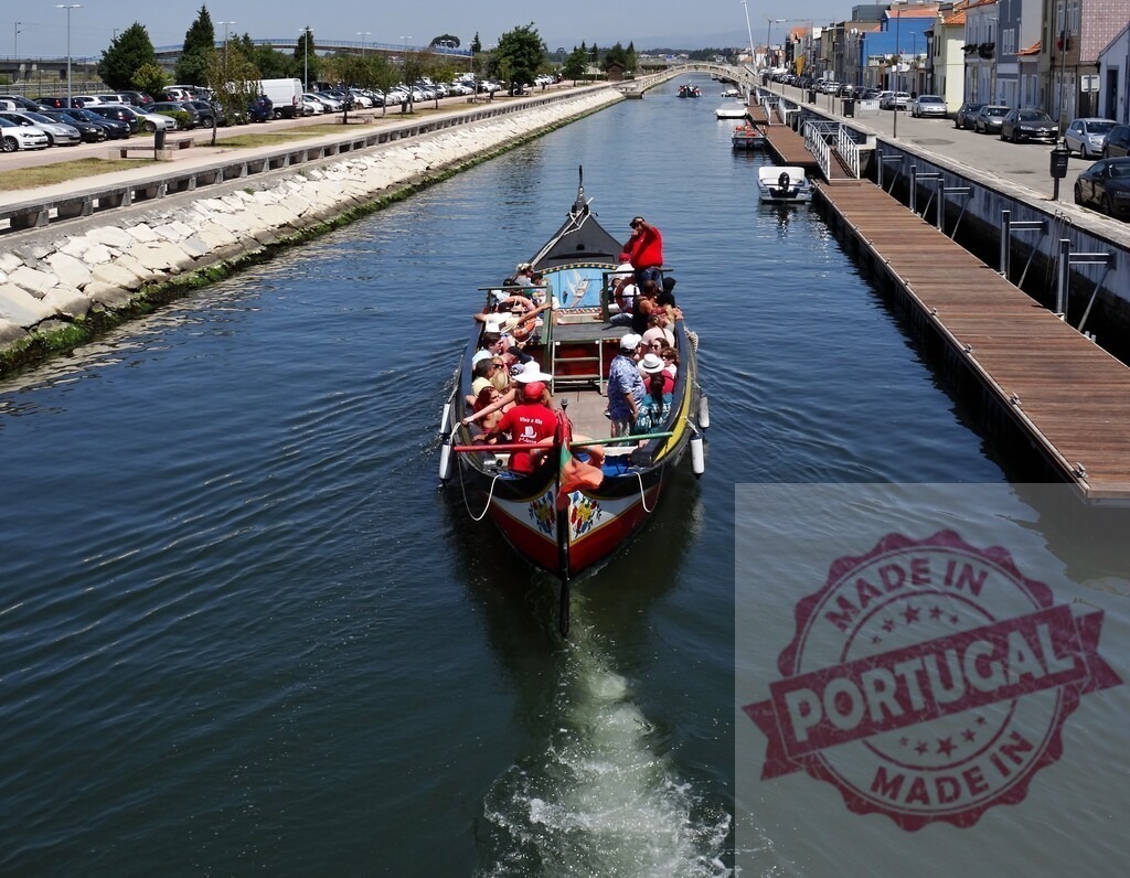 Aveiro moliceiro boat navigating a canal with tourists | Tourists enjoying a moliceiro boat ride along an Aveiro canal on a sunny day - Realisiert mit Pictrs.com