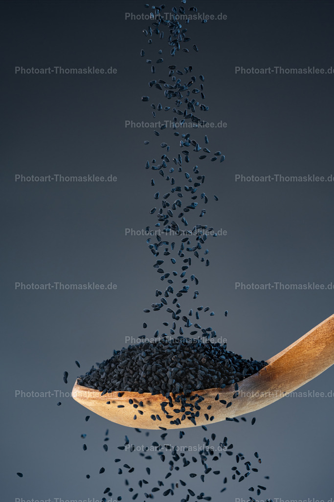 Gewürzregen - Magie des schwarzen Kümmels | Gewürzregen - Magie des schwarzen Kümmels. Kollektion: A spoon of tastefully seasoningsInhalt:Das Bild zeigt schwarzen Kümmel, der in Bewegung von oben herab auf einen Holzlöffel fällt. Die kleinen, mattschwarzen Samen schweben wie in Zeitlupe durch den Raum und sammeln sich in einem schlichten, naturfarbenen Löffel. Der Fokus liegt klar auf dem Gewürz selbst – jedes Samenkorn ist detailliert sichtbar und vermittelt Reinheit und Ursprünglichkeit.Technik:Die Fotografie nutzt eine Hochgeschwindigkeitsaufnahme mit starker Tiefenschärfe. Die Bewegung der fallenden Kümmelsamen wurde punktgenau eingefroren, was dem Bild Dynamik und Leichtigkeit verleiht. Der Hintergrund ist ein gleichmäßiger, neutraler Grauton, der weder ablenkt noch mit dem Motiv konkurriert.Farben:    Hauptfarben: Tiefschwarz der Kümmelkörner    Kontrastierend: Warmes Braun des Holzlöffels    Hintergrund: Klares, ruhiges Mittelgrau für moderne ÄsthetikDarstellung:Die Inszenierung ist reduziert, aber äußerst wirkungsvoll. Die Komposition spielt mit dem Kontrast zwischen Schlichtheit und Detailfülle, Bewegung und Ruhe. Der minimalistische Stil spricht Liebhaber von hochwertiger Food-Fotografie ebenso an wie Designaffine.Geeigneter Ort zum Aufhängen:    Moderne Küchen mit natürlichem Materialkonzept    Bio-Läden oder Gewürzgeschäfte    Restaurant oder Bistro mit Fokus auf gesunde, orientalische oder vegane Küche    Kochstudios oder als Teil einer Gewürzserie in einer Galerie oder Wohnküche - Realisiert mit Pictrs.com