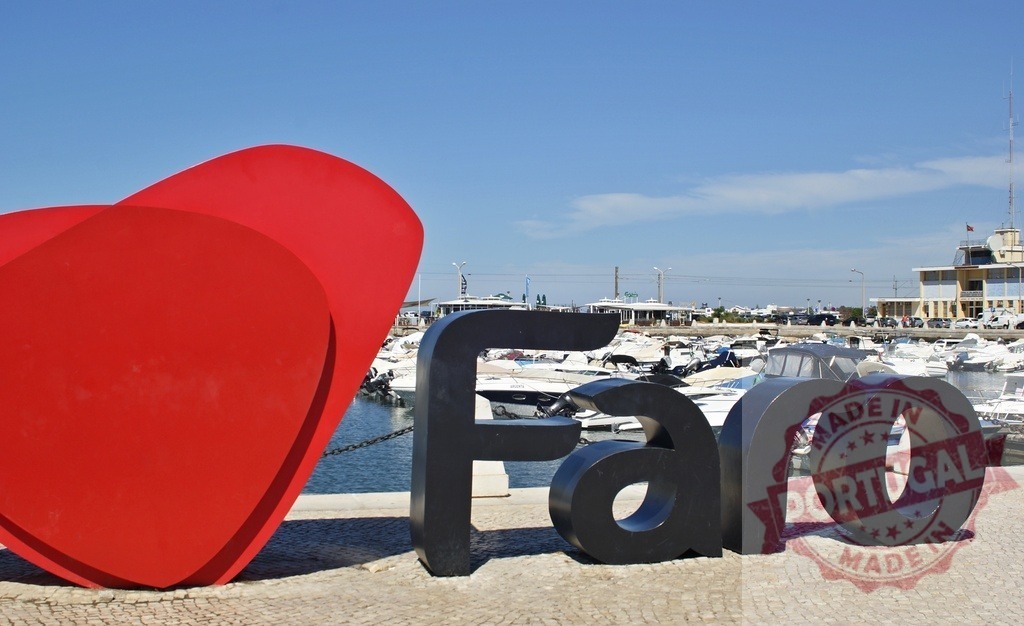 Faro sign with red heart sculpture welcoming tourists | Faro sign and heart sculpture standing by a marina filled with boats under a blue sky, representing travel - Realized with Pictrs.com