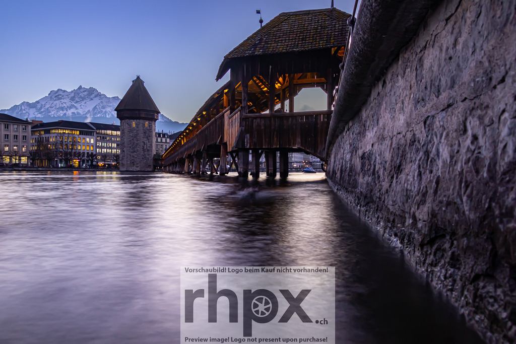 Abenddämmerung an der Kapellbrücke Luzern | Licht. Wasser. Historie.Die historische Kapellbrücke und der Wasserturm präsentieren sich in der blauen Stunde vor der markanten Kulisse des verschneiten Pilatus. Die sanfte Langzeitbelichtung glättet das Wasser der Reuss und lässt die warmen Lichter der Stadt besonders stimmungsvoll leuchten.Die Tiefenwirkung dieses Nachtmotivs kommt auf Acrylglas oder Alu-Dibond hervorragend zur Geltung. Wähle unter 'Produktauswahl' dein Wunschformat oder entdecke weitere Optionen wie Poster oder Downloads. - Realisiert mit Pictrs.com