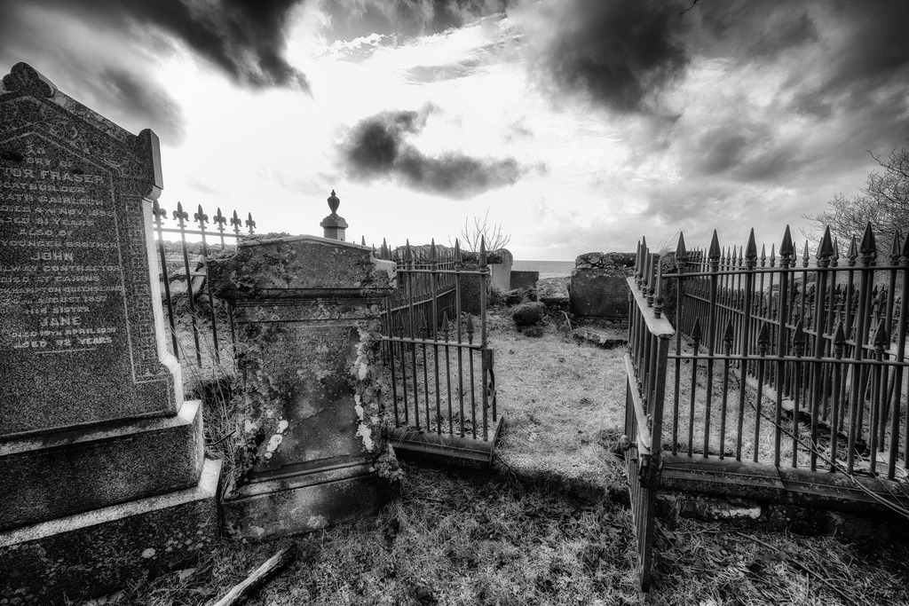 Friedhof mit Ausgang | Ein weitwinkliger Blick erfasst die melancholische Szenerie eines historischen Friedhofs an der Küste von Gairloch. Alte, verwitterte Grabsteine und schmiedeeiserne Zäune dominieren den Vordergrund, während ein dramatischer Himmel und das Meer den Horizont bilden. Die Schwarz-Weiss-Darstellung betont die Texturen und die zeitlose Atmosphäre des Ortes. - Realisiert mit Pictrs.com