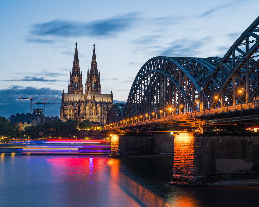 Lichtspuren an der Hohenzollernbrücke | Leuchtende Lichtspuren ziehen unter der Hohenzollernbrücke über den Rhein, während der Kölner Dom im Hintergrund die Stadtsilhouette prägt. Die Langzeitbelichtung verleiht der Szene eine dynamische Abendstimmung. - Realisiert mit Pictrs.com