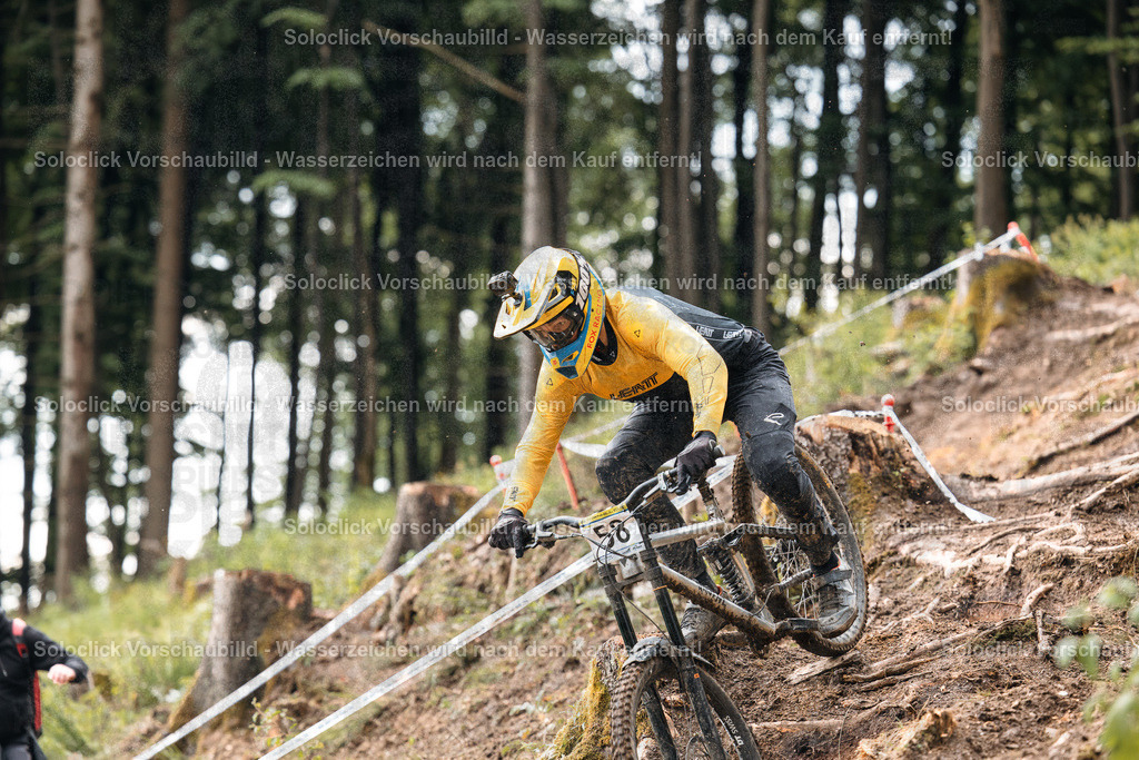 Mountainbike-Abenteuer | Ein Mountainbiker in voller Ausrüstung rast einen steilen, bewaldeten Hang hinunter. Er trägt einen gelben Helm und eine gelbe Brille, sowie schwarze Handschuhe und eine schwarze Hose. Er trägt die Nummer 58 auf seinem Rücken. Der Hintergrund besteht aus dichtem, grünen Laubwerk und einem schmalen, weißen Band, das den Kurs markiert. - Realisiert mit Pictrs.com