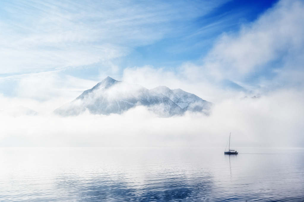 Thunersee: Segelboot vor Nebelbergen | Ein Segelboot gleitet ruhig über die glatte Oberfläche des Thunersees. Im Hintergrund erheben sich schneebedeckte Berge, teilweise in dichte Nebelschwaden gehüllt, unter einem klaren blauen Himmel. Die Komposition erzeugt eine Atmosphäre von Stille und majestätischer Natur. - Realisiert mit Pictrs.com