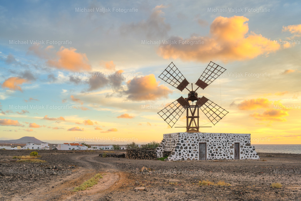 Windmühle in Puerto Lajas auf Fuerteventura | Bei Sonnenaufgang steht die Windmühle von Puerto Lajas still in der kühlen Morgenluft. Über ihr leuchten die Wolken bereits in kräftigem Orange, als hätte der Himmel den neuen Tag vorweggenommen. Die warmen Farben breiten sich über die Szene aus, während die Mühle dunkel und klar dagegensteht. Ein Moment voller Ruhe und Intensität zugleich. - Realisiert mit Pictrs.com