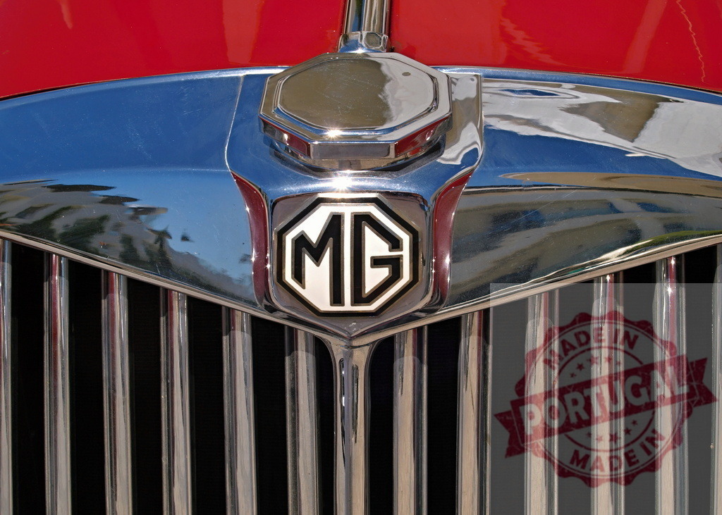 Shiny chrome MG radiator grill and logo on red vintage car body | Close-up of MG chrome radiator grill and logo on red vintage car body reflecting surrounding buildings - Realized with Pictrs.com