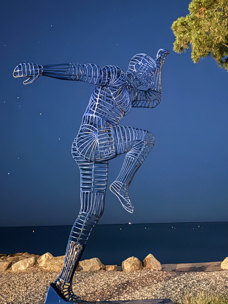 Illuminated Wire Runner Sculpture at Night | A captivating night photograph taken at the beach of Villeneuve-Loubet, France. The image features a modern, blue-illuminated wire sculpture of a runner in mid-stride against a deep night sky and the dark ocean. A powerful, dynamic piece of contemporary art, perfect as motivational wall decor for modern offices, sports facilities, or stylish homes. - Realisiert mit Pictrs.com