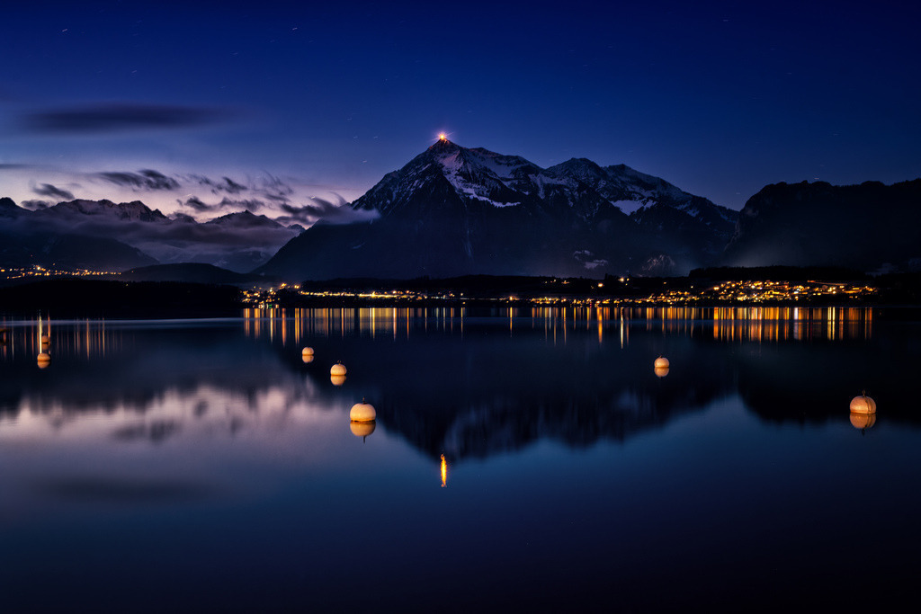 Niesen und Thunersee zur Blauen Stunde | Die Szenerie zeigt den Thunersee zur Blauen Stunde, mit dem markanten Niesen im Hintergrund, dessen Gipfel von einem Licht erhellt wird. Die Lichter der Uferbebauung spiegeln sich zusammen mit mehreren Bojen im ruhigen Wasser, während der Himmel erste Sterne zeigt. Diese Langzeitbelichtung betont die friedliche Atmosphäre und die klaren Reflexionen der alpinen Landschaft. - Realized with Pictrs.com
