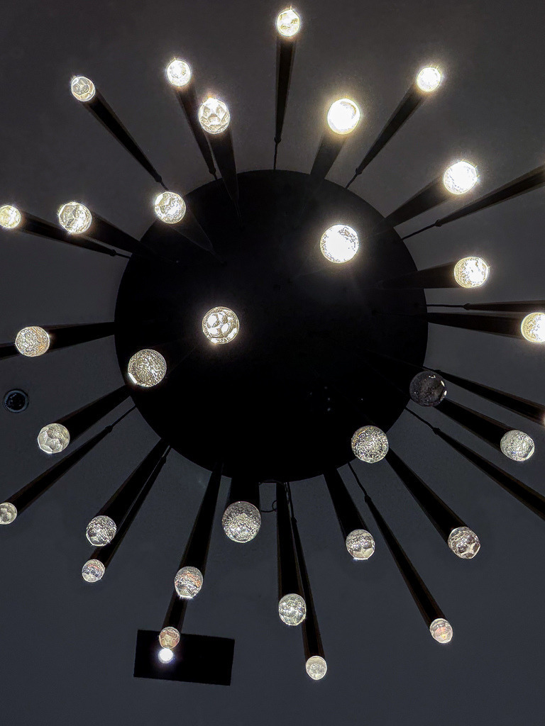 Lichtorbit – Abstraktes Galaxie Lichtkunst Wandbild | Ein faszinierender Mikrokosmos aus Licht und Schatten. Diese meisterhafte, perfekt zentrierte Fotokunst-Aufnahme fängt eine moderne Deckenleuchte aus der Froschperspektive ein. Die strahlenförmig angeordneten Lichtpunkte verschwimmen im harten Kontrast zur tiefen Dunkelheit zu einem hypnotischen, kosmischen Muster, das an eine ferne Galaxie erinnert. Ein hochexklusiver Design-Blickfang für minimalistische Wohnräume, urbane Lofts oder moderne Büros. Empfohlen als Premium-Druck unter Acrylglas für eine beeindruckende Tiefenwirkung. - Realisiert mit Pictrs.com
