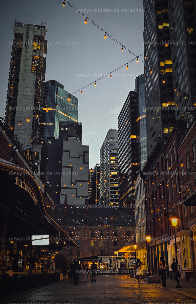 South Street Seaport zur blauen Stunde – Sommerabend voller Leben in Manhattan | Wenn die blaue Stunde über Manhattan hereinbricht, verwandelt sich das South Street Seaport in einen Ort voller Leben und Atmosphäre. Die historischen Straßen, die tagsüber ruhig wirken, füllen sich mit Menschen, die den Sommerabend genießen – beim Dinner in charmanten Restaurants, in lebhaften Bars oder beim entspannten Spaziergang entlang des Wassers. Die Lichter beginnen zu glühen, während der Himmel in tiefem Blau leuchtet und die Kulisse der Skyline den perfekten Hintergrund bildet. Es ist ein Moment, in dem Geschichte und Moderne verschmelzen und die einzigartige Energie New Yorks spürbar wird.