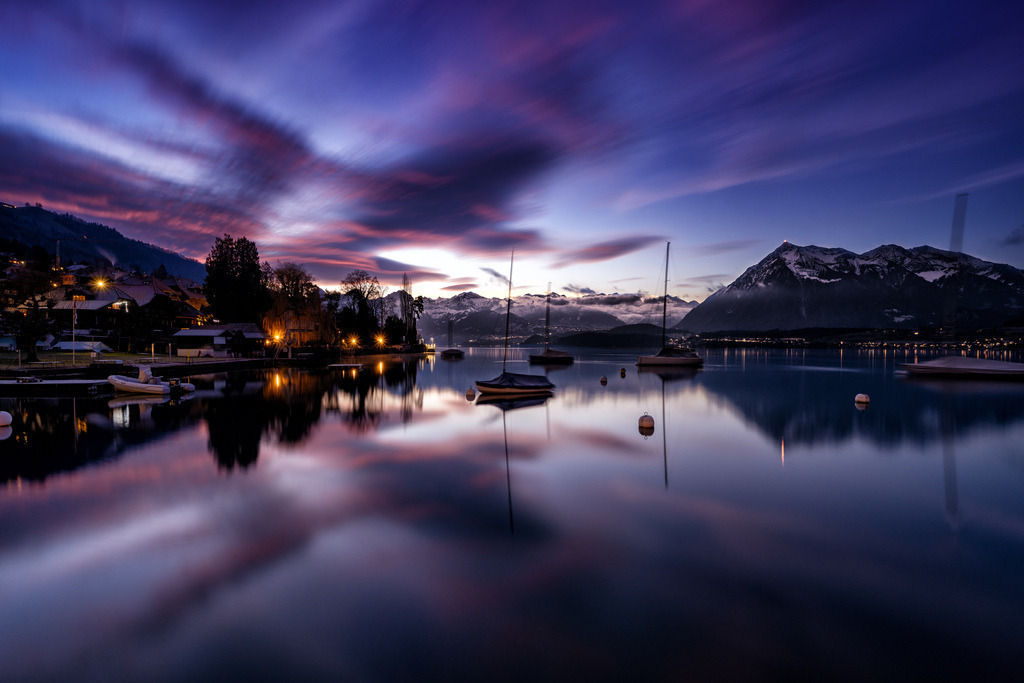 Morgenstimmung über dem Thunersee | Eine Langzeitbelichtung fängt die ruhige Morgenstimmung am Thunersee in Hilterfingen ein. Der Himmel zeigt dramatische purpur- und rosafarbene Wolkenstreifen, die sich perfekt im spiegelglatten Wasser widerspiegeln. Im Hintergrund erheben sich schneebedeckte Berge, während am Ufer die Lichter eines Dorfes leuchten. - Realisiert mit Pictrs.com