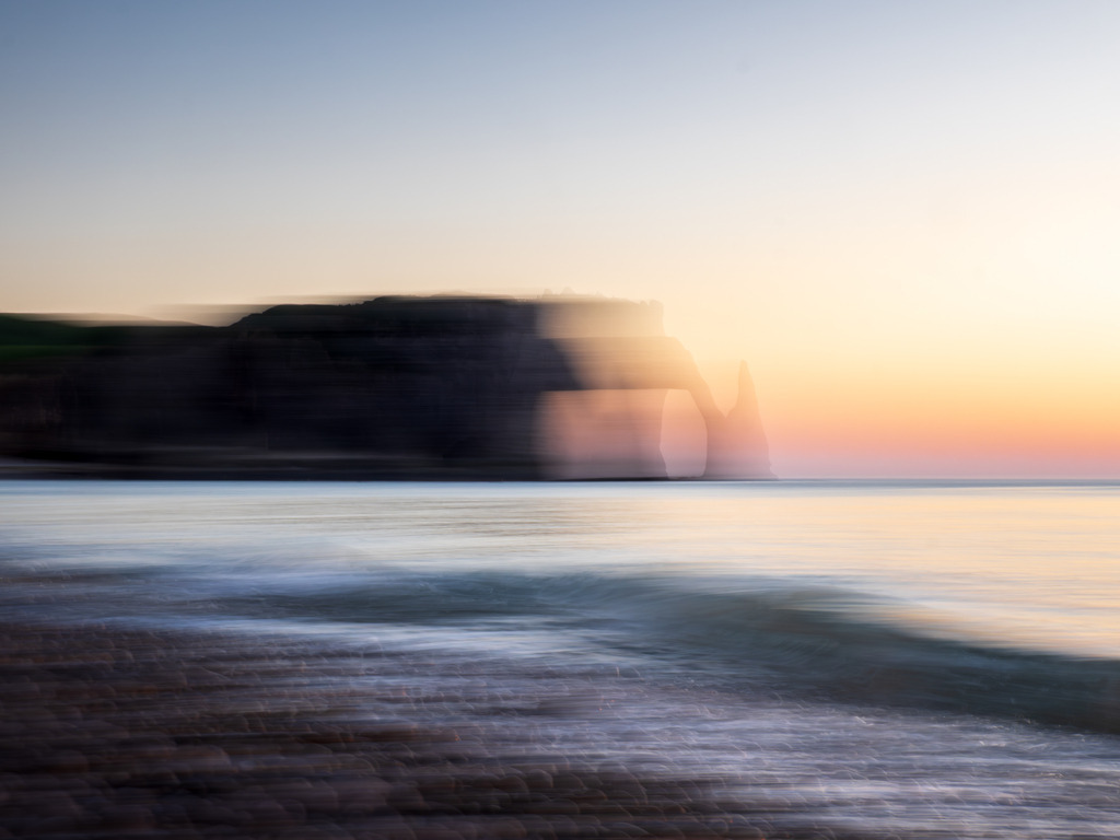 Gefährten des Lichts | Wie ein Echo aus einer anderen Welt liegt der Felsen von Étretat im Zwielicht – kaum greifbar, fast schon Erinnerung. Der Sonnenuntergang wirft goldene Schleier über das Meer, das wie flüssiges Glas am Ufer bricht. In der Spiegelung des Himmels scheint sich die Zeit selbst aufzulösen.Dies ist der Moment zwischen Atemzügen – ein Zwischenraum, in dem sich das Sichtbare entzieht und das Unsichtbare offenbart. Ein Ort, an dem sich das Licht nicht nur auf der Wasseroberfläche bricht, sondern tief im Innern etwas berührt. - Realized with Pictrs.com