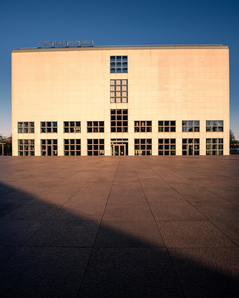 Moderne Architektur im Hamburger Abendlicht | Eine klare und minimalistische Architekturaufnahme in Hamburg. Das warme Licht auf der Fassade hebt die geometrischen Formen des Gebäudes hervor und bildet einen ruhigen Kontrast zum weitläufigen Vorplatz. - Realisiert mit Pictrs.com