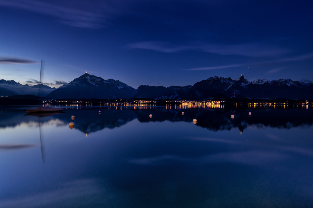 Thunersee, Hilterfingen: Blaue Stunde mit Niesen und Stockhorn. | Eine weite Ansicht des Thunersees zeigt die ruhige Wasseroberfläche, die die Lichter der Uferbebauung und die schneebedeckten Gipfel der Berner Alpen, darunter der beleuchtete Niesen, perfekt widerspiegelt. Die lange Belichtungszeit glättet das Wasser zu einer spiegelnden Fläche und betont die tiefblaue Stimmung der Dämmerung, während ein Segelboot am linken Bildrand sanft verschwommen erscheint. Die Komposition fängt die friedliche Atmosphäre der Schweizer Berglandschaft bei Nacht ein und bietet eine ästhetisch ansprechende Darstellung von Natur und menschlicher Zivilisation. - Realisiert mit Pictrs.com
