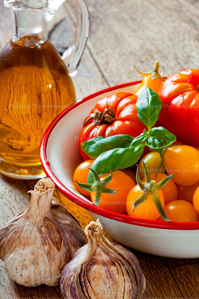 Bio Tomaten, Knoblauch und Olivenöl rustikalem Holz | Diese stimmungsvolle Küchenszene zeigt frische Tomaten in einer traditionellen Emailleschale mit Basilikumblättern und frischen Knoblauch. Große rote Tomaten liegen neben kleineren gelben Exemplaren, alle mit ihren natürlichen Stielen versehen. Eine bauchige Glasflasche enthält hochwertiges Olivenöl und ergänzt die mediterrane Atmosphäre perfekt. Die natürliche Holzmaserung des Untergrunds verleiht der Komposition Wärme und Authentizität für die anspruchsvolle Foodfotografie. - Realisiert mit Pictrs.com
