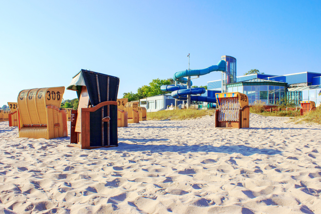 Wandbilder: Strandkörbe am Wellenbad in Eckernförde | Dieses Wandbild im Querformat zeigt Strandkörbe am Strand in Eckernförde. Im Hintergrund ist das Wellenbad zu sehen. Der blaue Himmel ist wolkenlos.  - Realisiert mit Pictrs.com