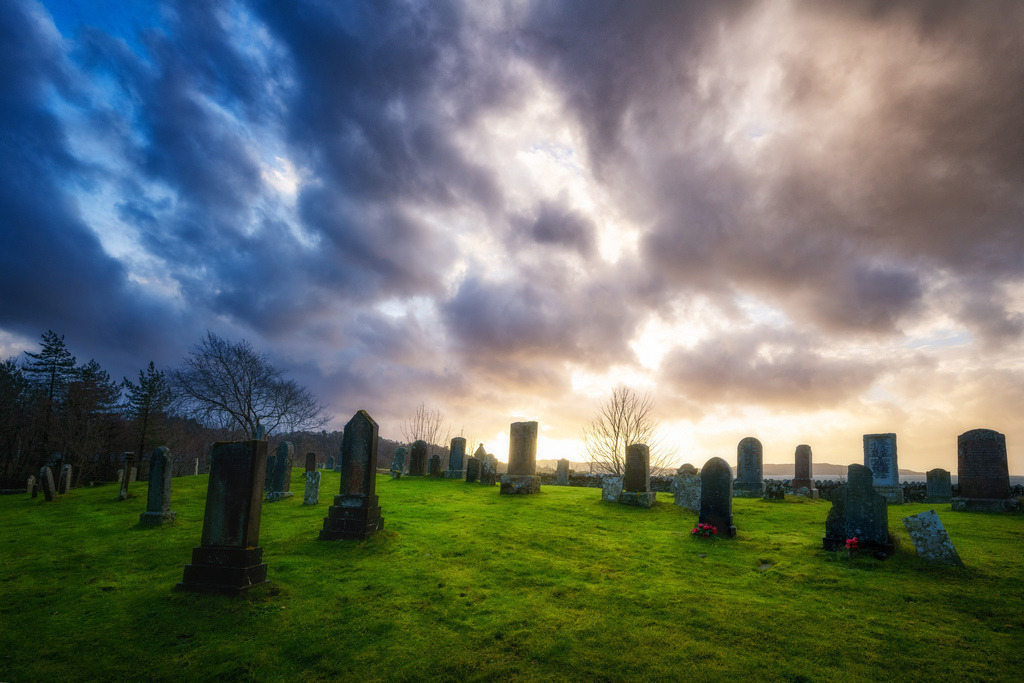 Liegen am Strand | Ein alter Friedhof in Gairloch, Schottland, präsentiert sich mit einer Reihe historischer Grabsteine auf einem grünen Hügel. Der Himmel zeigt eine beeindruckende Farbpalette von tiefem Blau und Grau auf der linken Seite bis zu warmen Goldtönen und hellen Wolken am Horizont, was eine starke visuelle Dynamik erzeugt. Die tief stehende Sonne beleuchtet die Szene von rechts und hebt die Silhouetten der Grabsteine und Bäume hervor. - Realisiert mit Pictrs.com