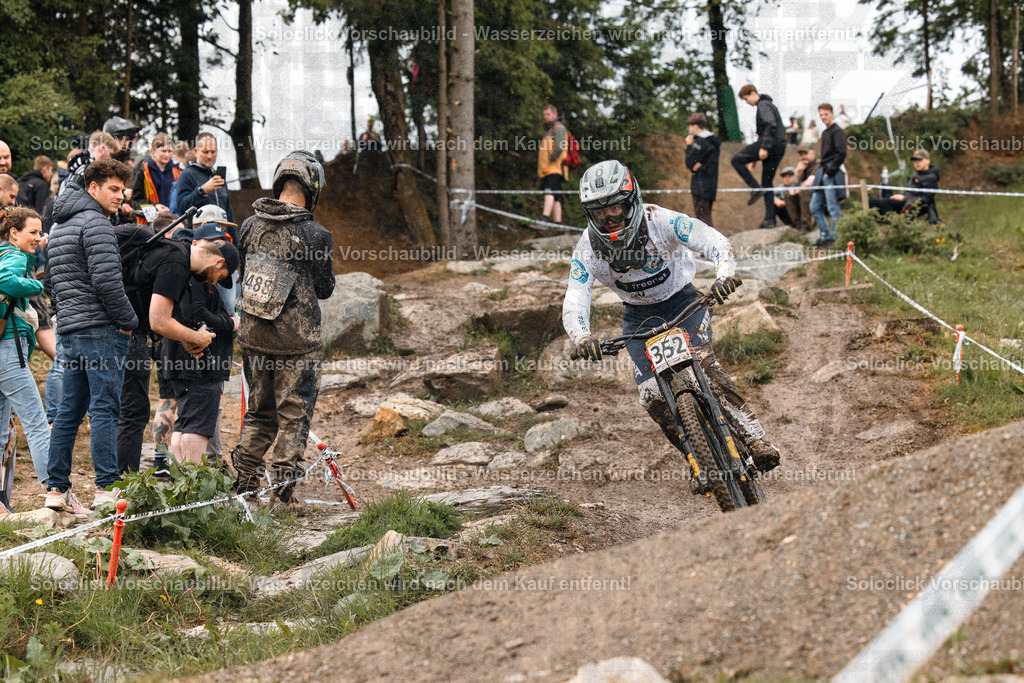 Mountainbike-Rennen im Wald | Das Bild zeigt einen Mountainbiker in voller Montur, der einen staubigen, steinigen Pfad mit hoher Geschwindigkeit hinunterfährt. Er trägt einen Helm, eine Schutzbrille und eine Weste mit der Nummer 352. Am Rand des Pfades stehen zahlreiche Zuschauer, die das Rennen beobachten. Der Pfad schlängelt sich durch eine bewaldete Landschaft. - Realisiert mit Pictrs.com