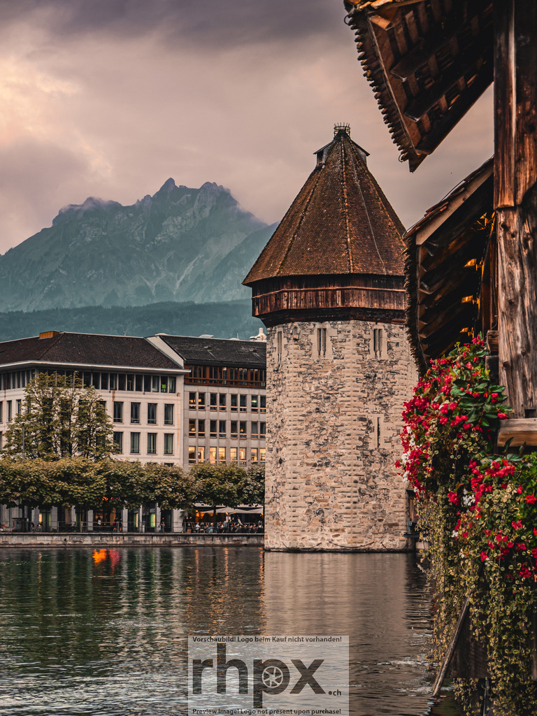 Wasserturm und Pilatus in Luzern | Geschichte. Stein. Gipfel.Der historische Wasserturm steht als stolzes Wahrzeichen inmitten der Reuss, während der markante Pilatus majestätisch über der Stadt wacht. Die sanfte Abendstimmung und die blühenden Details der Kapellbrücke verleihen dieser klassischen Luzerner Ansicht eine besondere Ruhe und Tiefe.Wähle unter "Produktauswahl" dein Wunschformat: Vom klassischen Wandbild über Puzzle &amp; Tassen bis zum digitalen Download (z.B. als Handy-Hintergrund). - Realisiert mit Pictrs.com