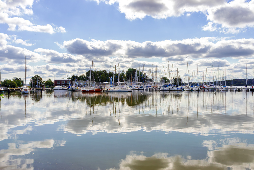 Wandbild: Spiegelung auf der Schlei in Schleswig | Dieses Wandbild im Querformat zeigt den Yachthafen in Schleswig. Auf der Schlei ist eine schöne Spiegelung zu sehen. Am blauen Himmel befinden sich einige sommerliche Wolken.  - Realisiert mit Pictrs.com