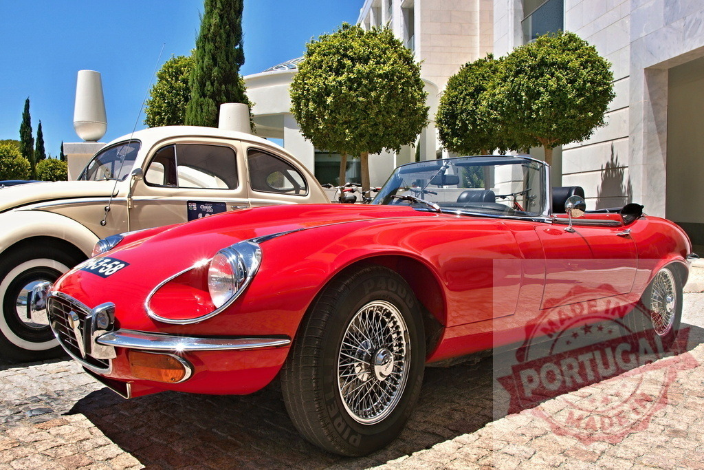 Red Jaguar E-Type parked near a Volkswagen Beetle in a luxury resort | Classic red Jaguar E-Type roadster parked near a vintage Volkswagen Beetle in front of a luxury hotel - Realized with Pictrs.com