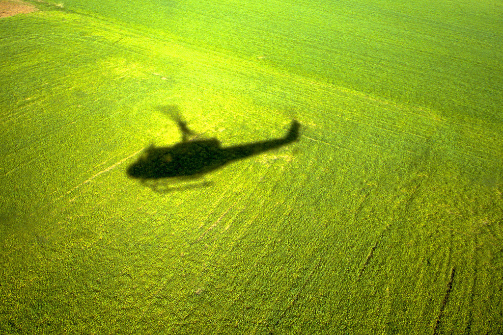 Bell UH1-D - Schatten | Die Aufnahme fängt den Schattenwurf eines Hubschraubers (typische Silhouette einer Bell UH-1D) ein, der sich scharf auf einem intensiv grünen Feld abzeichnet. Die Perspektive von oben lässt die Struktur der Vegetation fast wie einen Teppich wirken, während der dunkle Schatten eine starke Dynamik und Tiefe erzeugt. Das helle Sonnenlicht sorgt für einen hohen Kontrast und betont die Weite der Landschaft. (Dia Scan) - Realisiert mit Pictrs.com