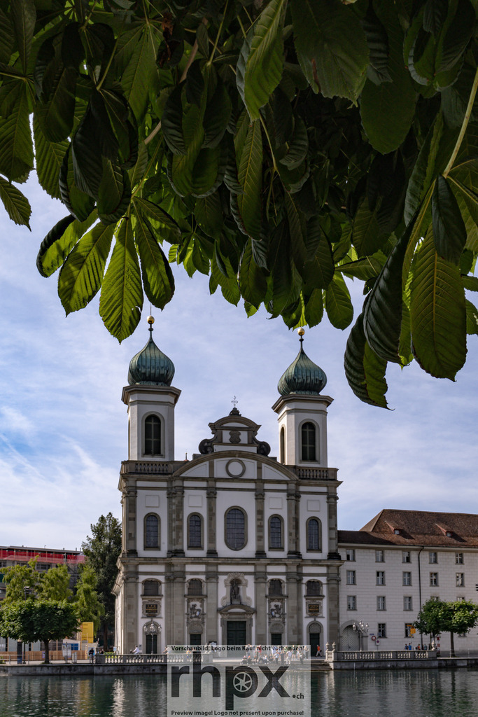 Jesuitenkirche Luzern an der Reuss | Aufnahme der Jesuitenkirche Luzern mit Blick von der Reuss. Die barocke Fassade wird von Kastanienblättern im Vordergrund eingerahmt, der Himmel zeigt leicht bewölktes Sommerlicht.  - Realisiert mit Pictrs.com