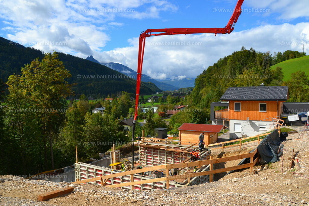 Betonieren von Betonwänden | Betonierarbeiten mit Betonpumpe auf Baustelle in Hanglage, dahinter Berglandschaft mit blauem Himmel und Wolken