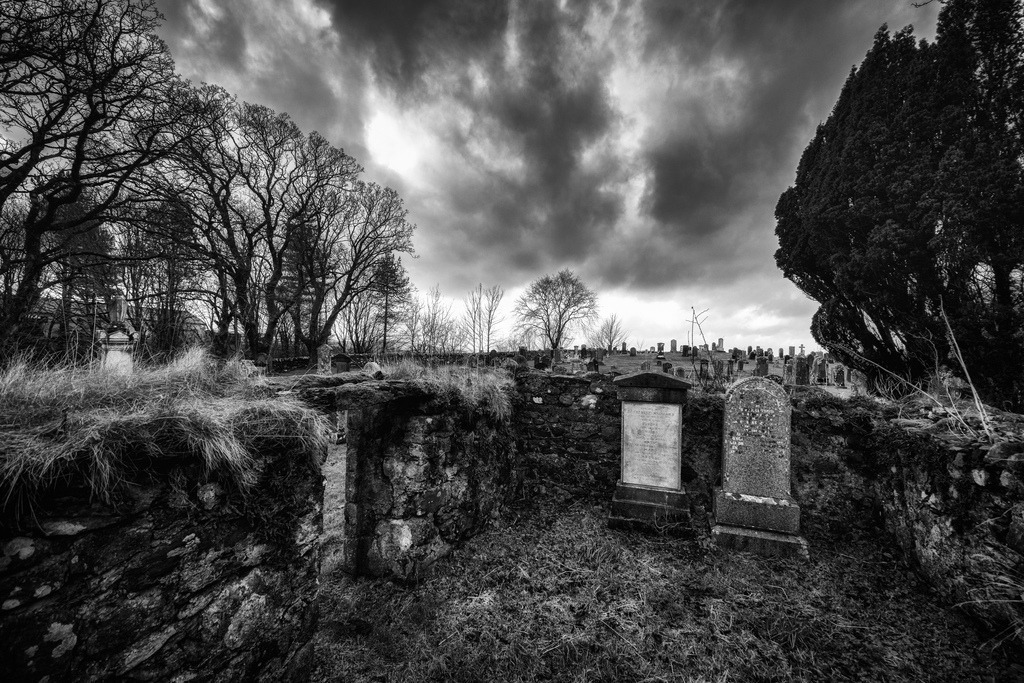 Chambre Séparée | Ein alter Friedhof in Gairloch, Schottland, praesentiert sich in einer Schwarz-Weiss-Aufnahme. Die Komposition wird von kahlen Baeumen, verwitterten Grabsteinen und einer alten Steinmauer dominiert. Ein dramatischer, wolkenverhangener Himmel bildet den oberen Abschluss und verleiht der Szene eine melancholische Stimmung. - Realisiert mit Pictrs.com