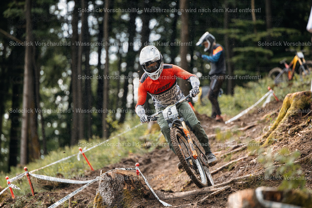 Mountainbike-Action im Wald | Das Bild zeigt einen Mountainbiker in voller Montur, der einen steilen, bewaldeten Hang hinabfährt. Er ist in einer dynamischen Position, die auf hohe Geschwindigkeit und Kontrolle hindeutet. Der Hintergrund besteht aus dichtem Wald, der die Herausforderung des Geländes unterstreicht. - Realisiert mit Pictrs.com