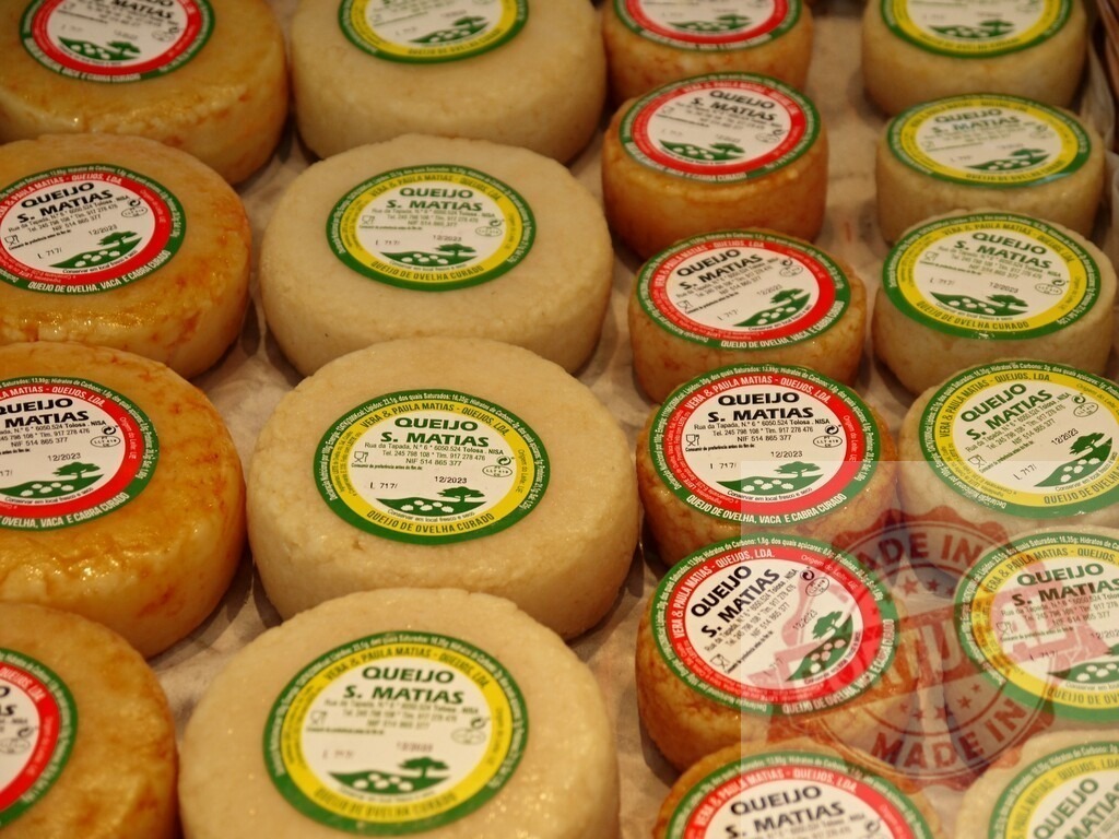 Queijo S. Matias cheese wheels on market stall | Artisanal Portuguese Queijo S. Matias cheese wheels displayed for sale, showing red and green labels - Realized with Pictrs.com