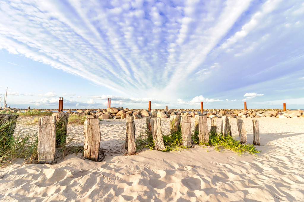 Wandbild: Holzpfähle am Sandstrand | Die beruhigende Kraft der Küstenlandschaft – dieses Wandbild zeigt die natürliche Schönheit des Strandes mit feinem Sand, einem sanften Sandfang und einer Steinmole im Hintergrund. Die Wolkenformation am Himmel schafft eine offene und entspannte Bildwirkung. Ideal für Wartezimmer, Behandlungsräume oder Empfangsbereiche, um eine stressfreie und wohltuende Umgebung zu gestalten. - Realisiert mit Pictrs.com
