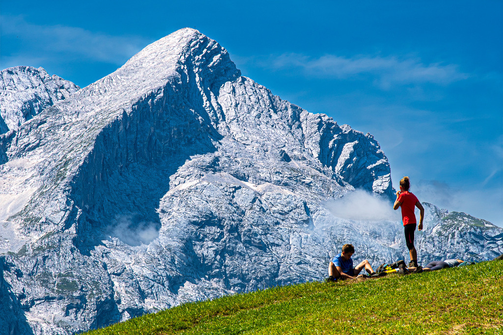 Gipfelglück und Rast an der Alpspitze | Vor der imposanten, schroffen Felskulisse der Alpspitze genießen Wanderer eine wohlverdiente Pause auf einer saftig grünen Bergwiese. Die erhabene Pyramide des Alpspitz-Gipfels ragt majestätisch in den strahlend blauen Sommerhimmel, während zarte Schleierwolken die Felswände umspielen. Das Bild fängt die perfekte Balance aus sportlicher Herausforderung und friedlicher Entspannung im Werdenfelser Land nahe Garmisch-Partenkirchen ein – ein Moment pürer Freiheit und alpiner Sommeridylle. - Realisiert mit Pictrs.com