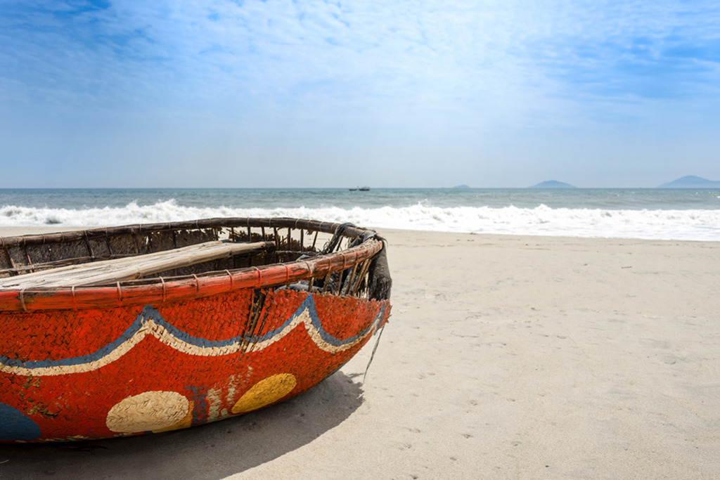 Traditionelles Korbboot | Vietnam | Premium-Wandbild von justsomewhere.com: Ein farbenfroh in Rot, Blau und Gelb bemaltes, traditionelles vietnamesisches Korbboot (Thuyền thúng) ruht auf dem hellen Sand eines Strandes in Vietnam vor der Kulisse brechender Meereswellen unter blauem Himmel. - Realisiert mit Pictrs.com