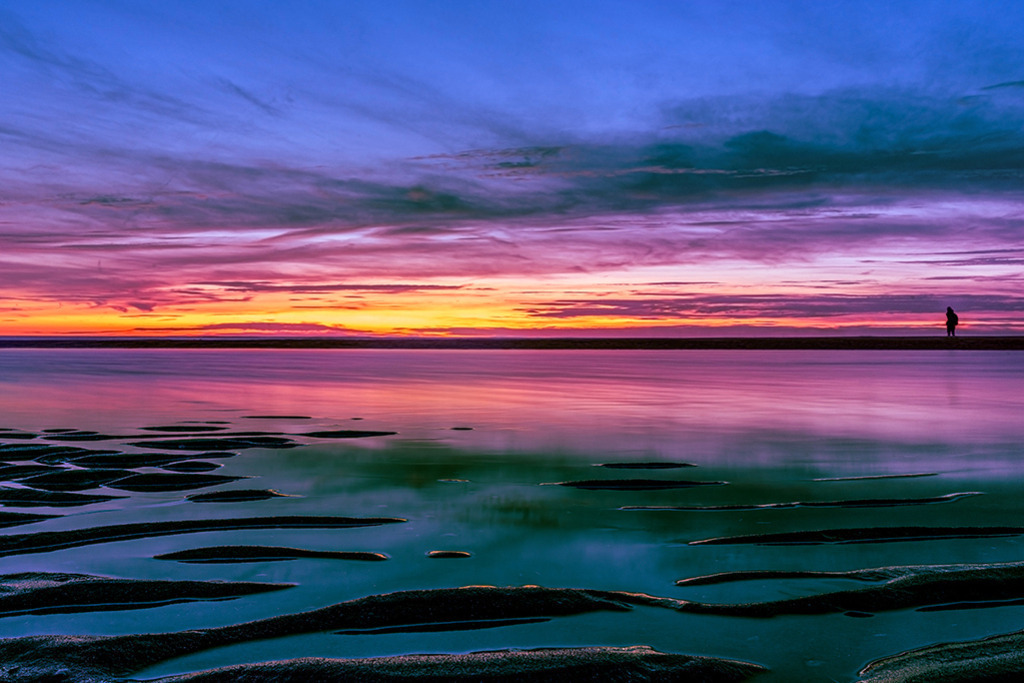 Der Wanderer im Wattenmeer | Deutschland | Premium-Wandbild von justsomewhere.com: Eine atemberaubende, farbintensive Landschaftsaufnahme des deutschen Wattenmeeres bei Sonnenuntergang mit einem violett-orangefarbenen Himmel, grünlichen Lichtreflexen im geriffelten Sand und der fernen Silhouette eines einzelnen Wanderers am Horizont. - Realisiert mit Pictrs.com