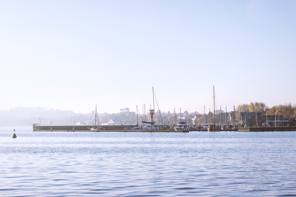 Wandbild: Yachthafen Eckernförde in morgendlicher Lichtstimmung | Der Blick über das stille Hafenwasser von Eckernförde zeigt Segelboote, Masten und die sanfte Weite der Küste im klaren Licht. Die ruhigen Wellen, die feinen Spiegelungen und die harmonische Linie der Boote schaffen eine Atmosphäre von Gelassenheit und maritimer Eleganz. Ein Motiv, das die friedliche Seite des Hafens und die besondere Stimmung der Ostseeküste eindrucksvoll einfängt.