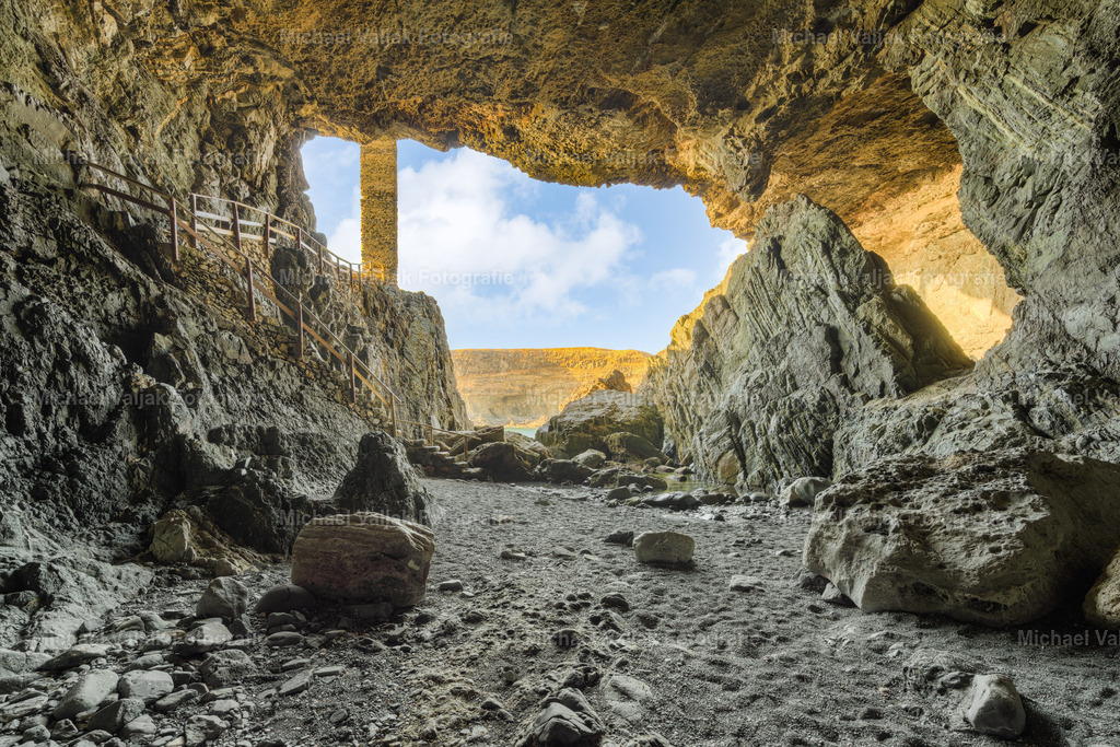 Höhlen von Ajuy auf Fuerteventura | Aus dem Inneren der Höhle von Ajuy fällt der Blick hinaus auf die hellen, sonnengebleichten Felsen der Westküste. Der dunkle Innenraum rahmt die Szene wie ein natürlicher Kontrast, während draußen das warme Licht die Schichten des Gesteins klar hervorhebt. Die Formen der Felswände wirken ruhig und kraftvoll zugleich, und der Übergang von tiefem Schatten zu strahlender Helligkeit verleiht dem Moment eine besondere Tiefe. - Realisiert mit Pictrs.com